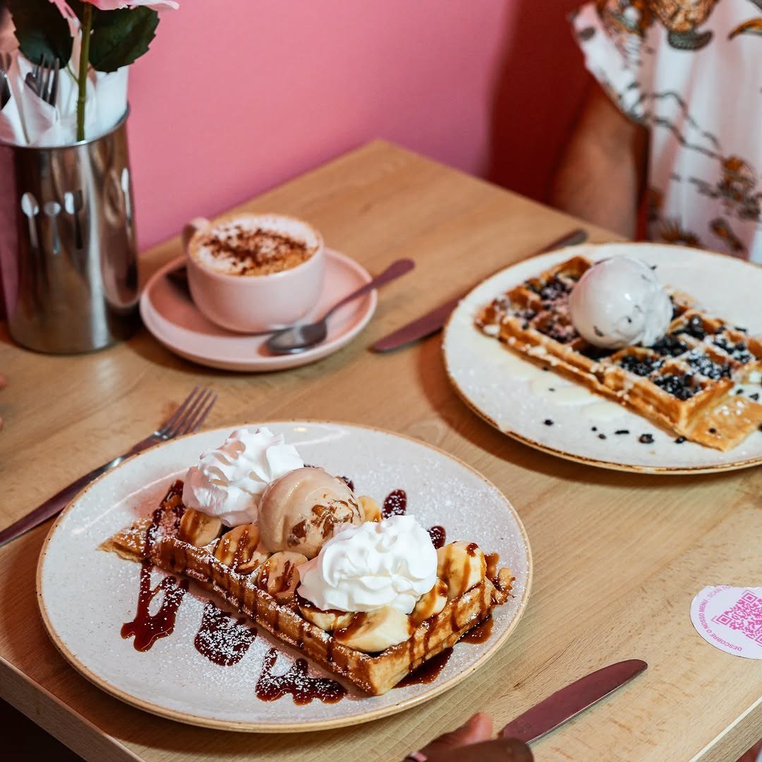 Mesa de madeira com waffles, gelado, natas e uma chávena, encostada a uma parede cor-de-rosa
