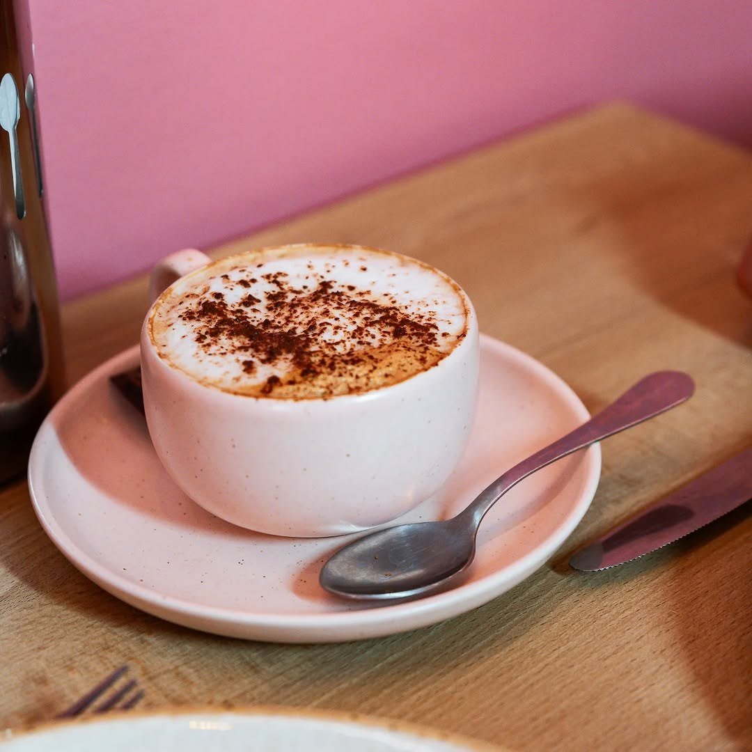 Chávena de cappuccino com cacau polvilhado numa mesa de madeira, junto a uma parede cor-de-rosa